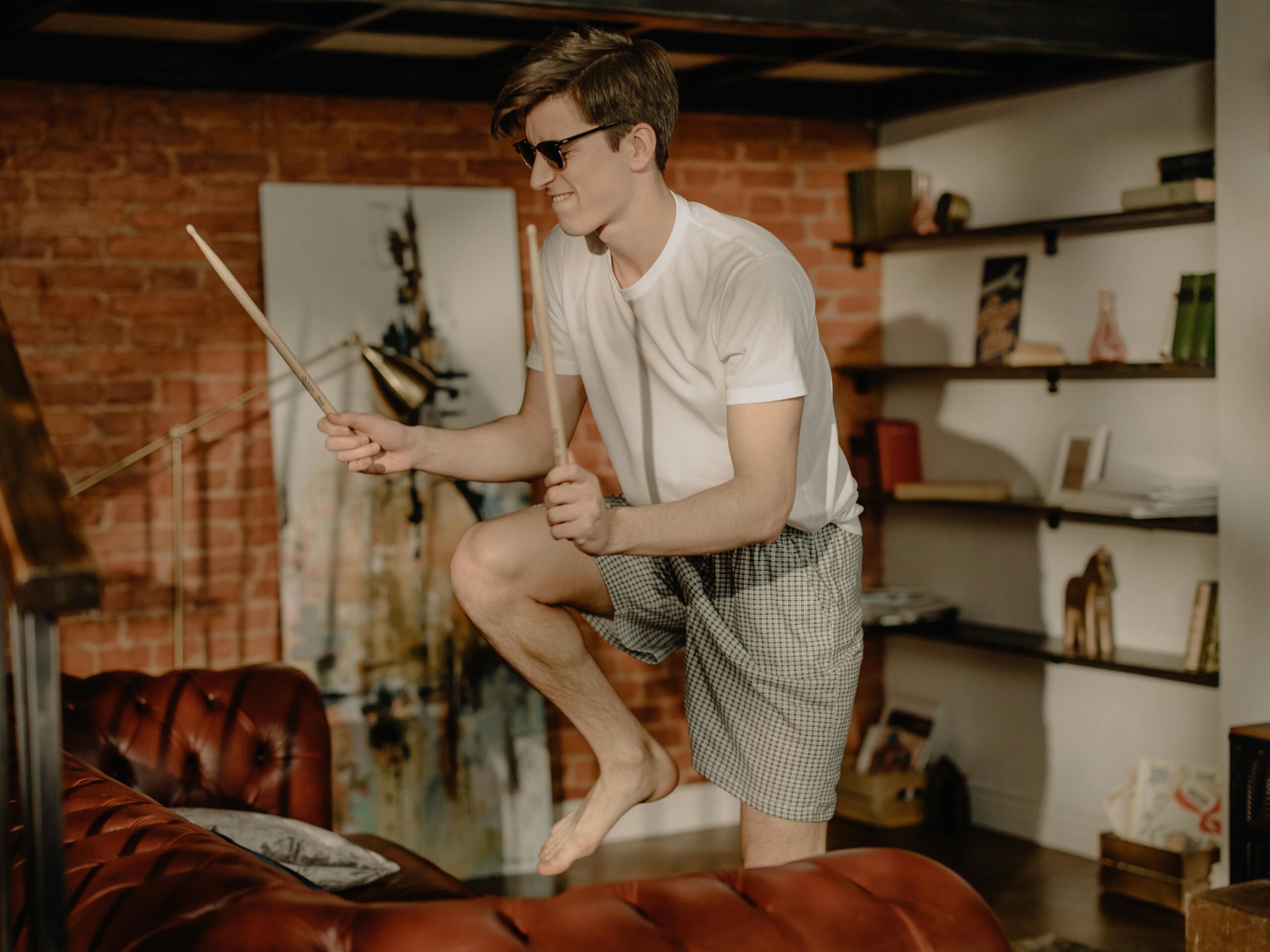Young man wearing sunglasses drumming with sticks in a cozy home setting.